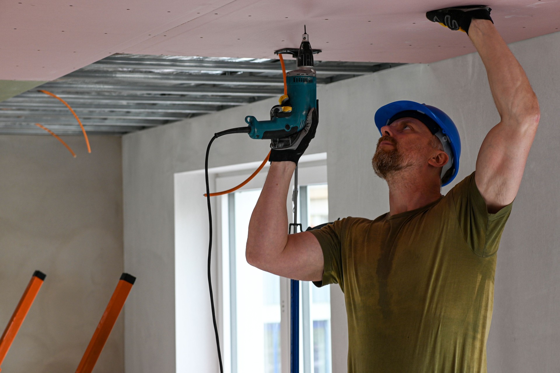 Construction worker wearing protective gear using power drill to install drywall on ceiling in new home construction